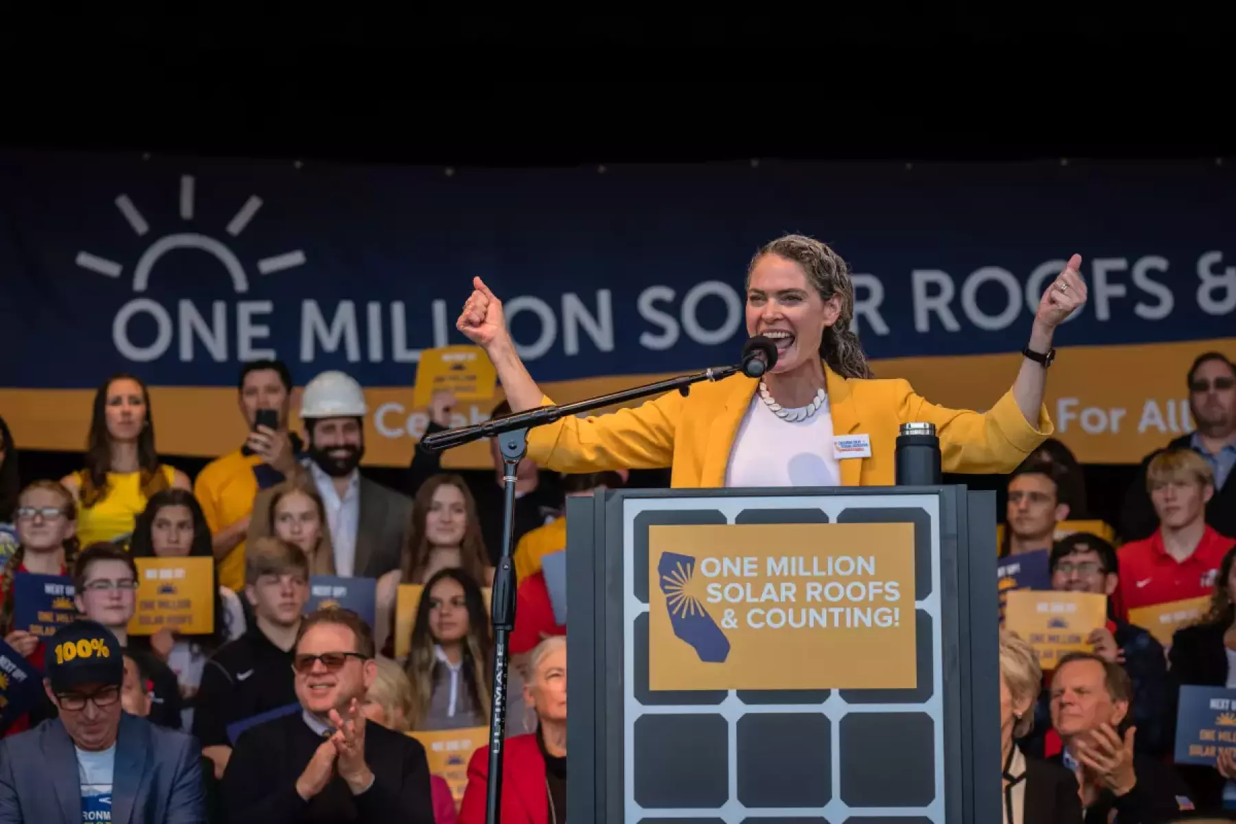 A woman raises and spreads her arms at a lectern.